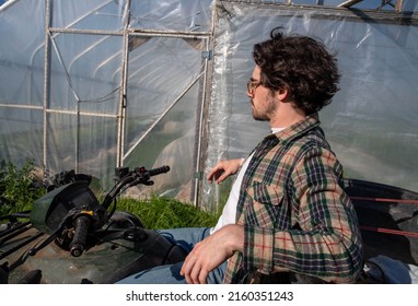 Young Caucasion Male Farmer On An Off Road Farm Vehicle Contemplating Beside An Organic Vegetable Garden Greenhouse In Natural Golden Hour Sunlight With Copy Space.