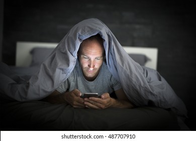 A Young Cell Phone Addict Man Awake Late At Night In Bed Using Smartphone