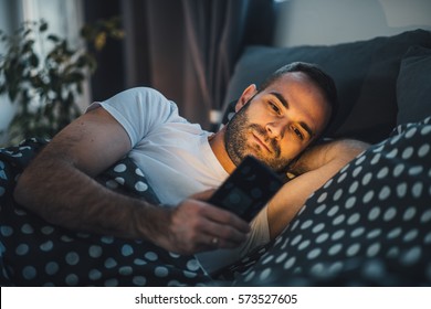 Young Cell Phone Addict Man Awake At Night In Bed Using Smartphone For Chatting, Flirting And Sending Text Message In Internet Addiction And Mobile Abuse Concept