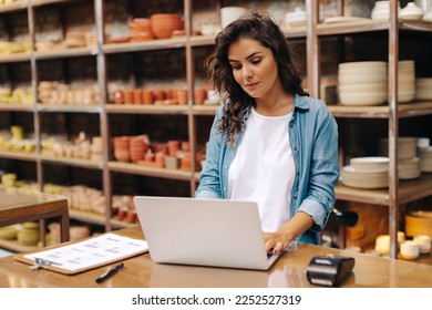 Young ceramic shop owner using a laptop while working in her store. Female entrepreneur managing online orders on her website. Confident young businesswoman running a creative small business. - Powered by Shutterstock