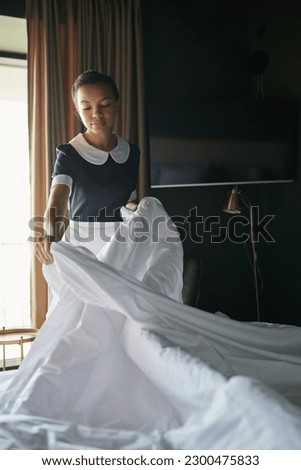 Young chambermaid in uniform standing by double bed in hotel room and changing blanket and other bedclothes before arrival of new guests