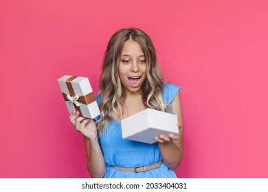 A Young Charmig Amazed Excited Blonde Woman With Wavy Hair In A Blue Dress Is Surprised Looking Into An Open Gift Box Isolated On A Color Pink Background. A Satisfied Girl Received A Pleasant Present