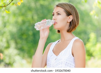 Young Charming Calm Woman In A White Blouse Drink Water From A Bottle In A Summer Green Park.