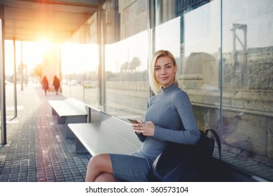 Young Charming Caucasian Female With Trendy Look Use Mobile Phone While Waiting Taxi On Bus Stop, Woman Holding Cell Telephone While Sitting On A Station Against Sunset Background With Copy Space     