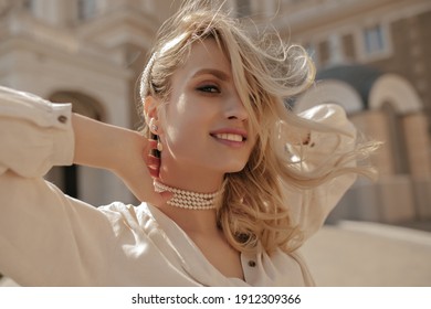 Young charming curly blonde woman in stylish white blouse and pearl necklace smiles sincerely and walks outside.