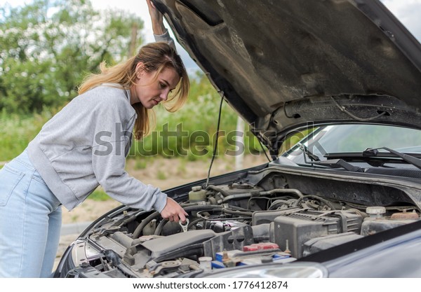 Young charming girl stands near the open hood\
of a broken car trying to repair\
it