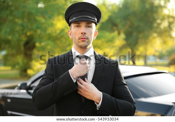 Young chauffeur adjusting tie near luxury car on\
the street