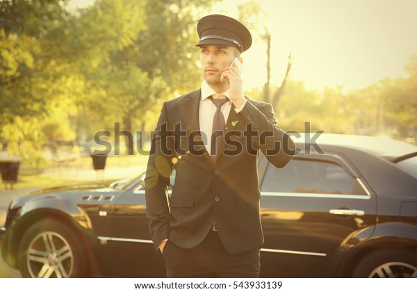 Young chauffeur speaking by cellphone near luxury\
car on the street