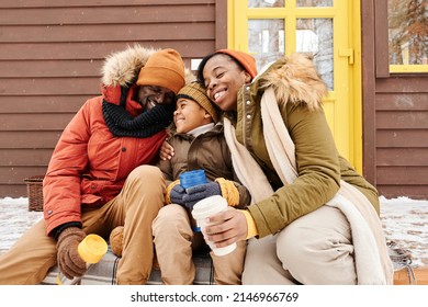Young cheerful and affectionate African American family of three with cups having tea by country house while sitting on porch