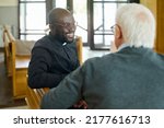 Young cheerful African American pastor of evangelical church consulting aged grey haired male parishioner after sermon