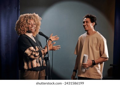 Young cheerful African American woman presenting audience new male comedian of stand up club and speaking in microphone - Powered by Shutterstock