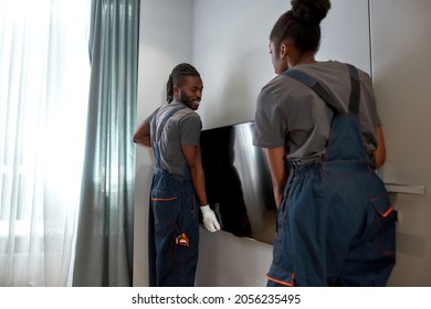 Young Cheerful African Handy Man And Woman Holding Large Flat Screen Tv, Setting It Onto Wall. Workpersons In Overalls Installing Television In New House.