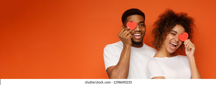 Young cheerful african-american couple in love holding red hearts over eyes and smiling, orange panorama background with empty space