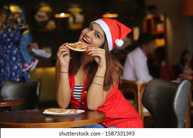 Young cheerful asian indian girl wearing santa hat eating pizza in a cafe restaurant, having fun with food. enjoying celebrating christmas holiday during covid-19 pandemic , lifestyle concept. 