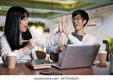 A young, cheerful Asian male college student is giving a high five to his friend and celebrating good news while doing homework together in a coffee shop. project winning, high score exam - Powered by Shutterstock