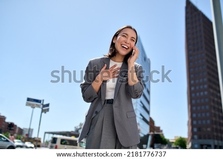 Young cheerful Asian successful businesswoman wearing suit standing on city street talking on mobile phone. Smiling woman making business call on cell feeling happy about good news outdoors.