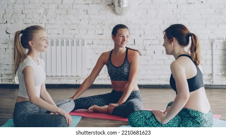 Young Cheerful Attractive Women Are Socialising After Yoga Practice Sitting On Mats In Circle In Light Studio. They Are Talking Emotionally, Laughing, Gesturing. Wellness Fun Concept.