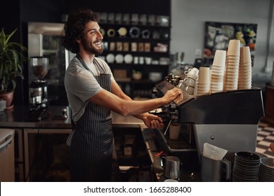 Young cheerful barista wearing black apron while preparing coffee at an automatic machine in a modern coffee shop