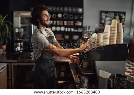 Young cheerful barista wearing black apron while preparing coffee at an automatic machine in a modern coffee shop