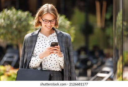 Young cheerful business woman in stylish suit standing outside in sunny day and looking with toothy smile at screen of modern smartphone in hand, chatting with friend or writing message to colleague - Powered by Shutterstock