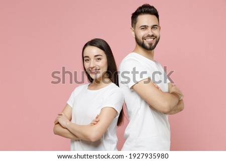 Young cheerful confident couple two friends man woman in white basic blank t-shirts standing bacl to back with folded crossed hands look camera isolated on pastel pink color background studio portrait