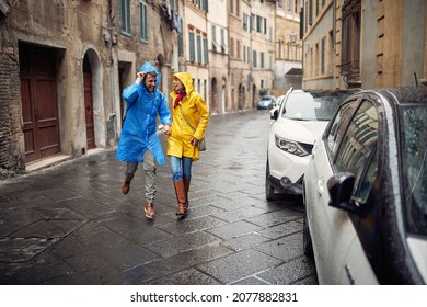 A Young Cheerful Couple In Raincoats Is Having Fun While Running From The Rain Towards The Car During A Walk The City On A Cloudy Day. Walk, Rain, City, Relationship