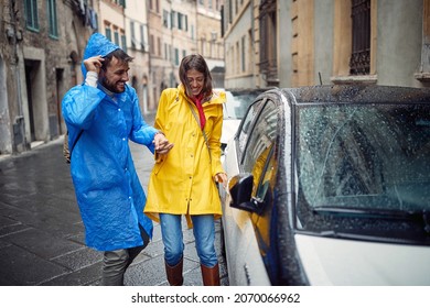 A Young Cheerful Couple In Raincoats Reached The Car While Running Away From The Rain On A Cloudy Day In The City. Walk, Rain, City, Relationship