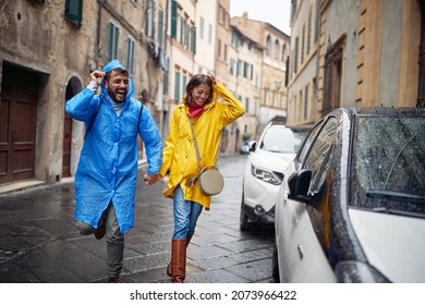 A Young Cheerful Couple In Raincoats Is Running From The Rain Towards The Car While Took A Walk The City In A Rainy Atmosphere. Walk, Rain, City, Relationship