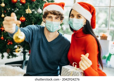 Young cheerful couple in Santa hats and wearing medical masks in quarantine sitting surround surrounded by colorful gift boxes enjoying celebrating Christmas at home.