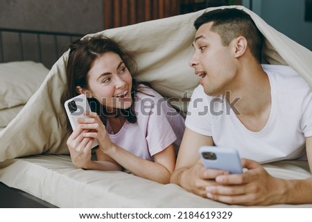 Young cheerful couple two family man woman she he wear t-shirt pajama lying in bed under blanket use mobile cell phone peep spend time together in bedroom home in own room hotel Real estate concept