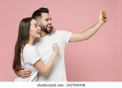 Young cheerful couple two friends bearded man brunette woman in white basic blank print design t-shirts doing selfie shot on mobile cell phone isolated on pastel pink color background studio portrait