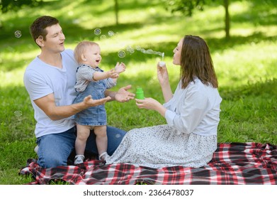 A young cheerful family with a baby is having a picnic in the park,blowing soap bubbles. Summer time concept.Family day