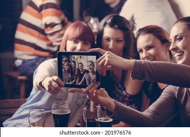 Young Cheerful Female Friends Taking Selfie In Pub Using Digital Tablet