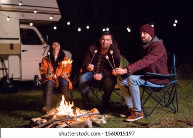 Young And Cheerful Friends Sitting Around Camp Fire And Roasting Marshmallows.