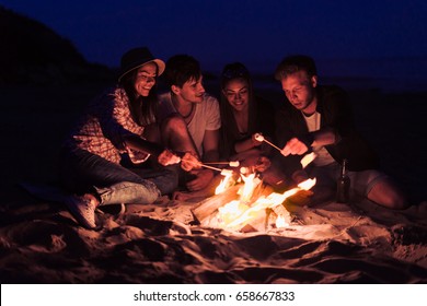 young and cheerful friends sitting on the beach and fry marshmallows near bonfire They look happy and smiling. Night time - Powered by Shutterstock