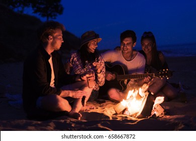 young and cheerful friends sitting on beach and fry sasuages or weenies in bonfire One man is playing guitar. Music on Wild beach. Happy couple relationships. - Powered by Shutterstock