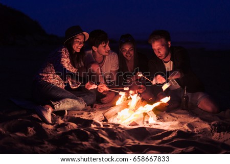 young and cheerful friends sitting on the beach and fry marshmallows near bonfire They look happy and smiling. Night time