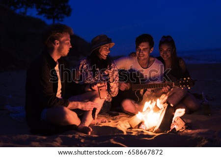 young and cheerful friends sitting on beach and fry sasuages or weenies in bonfire One man is playing guitar. Music on Wild beach. Happy couple relationships.