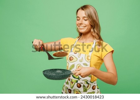 Young cheerful fun housewife housekeeper chef cook baker woman wear apron yellow t-shirt hold in hand pan spatula frying pancakes isolated on plain pastel green background studio. Cooking food concept