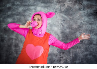 Young And Cheerful Girl In Colorful Pink Costume Of Pig With A Snout Instead Of A Nose On A Gray Background. April Fool's Day. Animator, Dancer Or Actor. Carnival, Show, Masquerade.