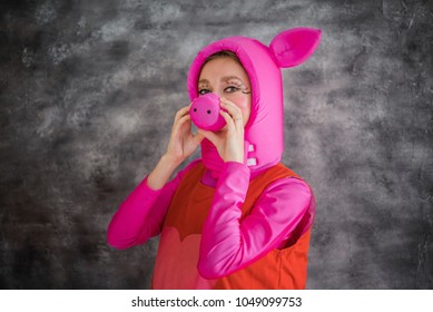 Young And Cheerful Girl In Colorful Pink Costume Of Pig With A Snout Instead Of A Nose On A Gray Background. Animator, Dancer Or Actor. Carnival, Show, Masquerade.