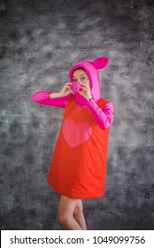 Young And Cheerful Girl In Colorful Pink Costume Of Pig With A Snout Instead Of A Nose On A Gray Background. Animator, Dancer Or Actor. Carnival, Show, Masquerade.