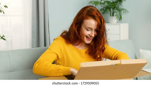 Young Cheerful Girl Getting Parcel At Home. Pretty Girl Sitting On Sofa At Home And Opening Carton Box. Mail Delivery To House.