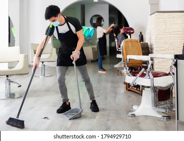 Young Cheerful  Glad Man Hairdresser In Mask Cleaning Floor In The Beauty Salon