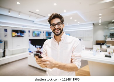 Young Cheerful Handsome Man In A White Shirt Is Using Phone In A Very Bright Tech Store.