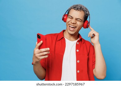 Young cheerful happy fun vivid man of African American ethnicity 20s he wearing red shirt headphones listen music sing song use mobile cell phone isolated on plain pastel light blue cyan background - Powered by Shutterstock