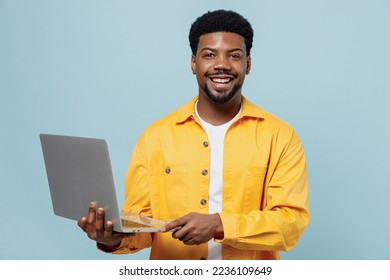 Young cheerful happy man of African American ethnicity 20s in yellow shirt hold use work on laptop pc computer isolated on plain pastel light blue background studio portrait. People lifestyle concept