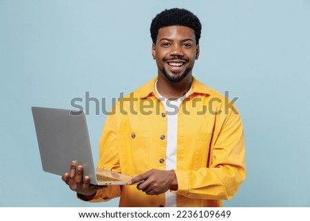 Young cheerful happy man of African American ethnicity 20s in yellow shirt hold use work on laptop pc computer isolated on plain pastel light blue background studio portrait. People lifestyle concept