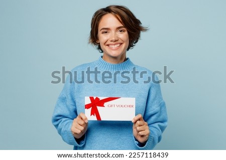 Young cheerful happy smiling woman wear knitted sweater hold gift certificate coupon voucher card for store isolated on plain pastel light blue cyan background studio portrait People lifestyle concept