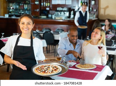 Young Cheerful Hispanic Woman Pizzeria Owner Inviting To Taste Delicious Pizza In Cozy Atmosphere